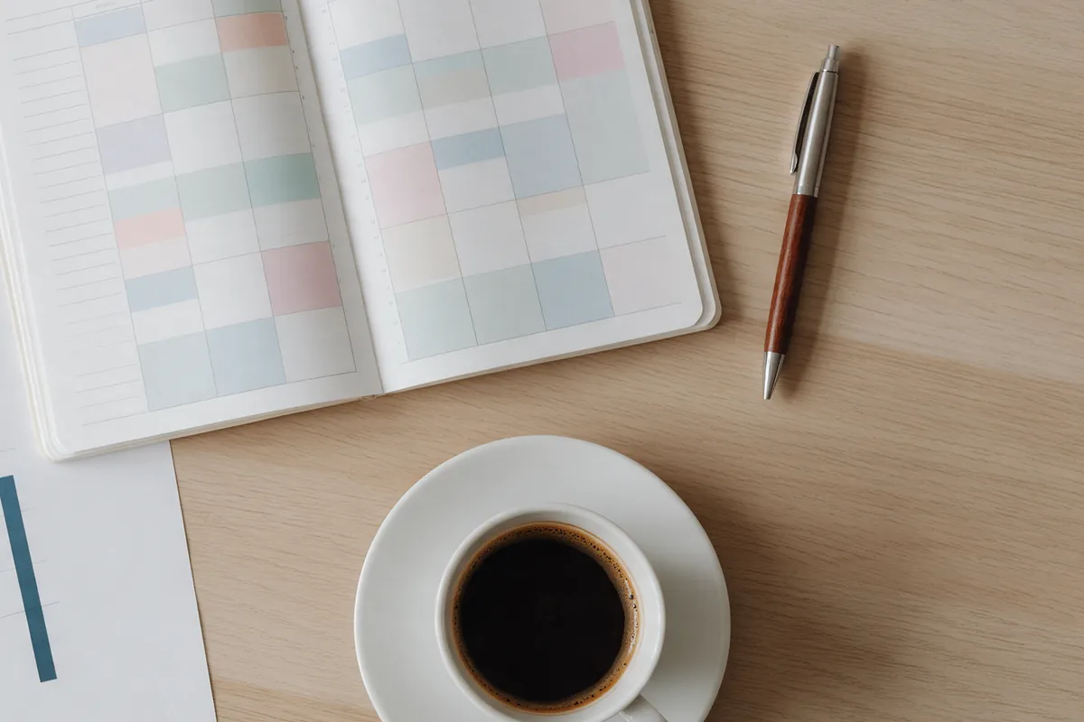 En utskriven rapport med stapeldiagram bredvid en öppen tidbok med färgade tider, en penna och en kaffe på ett ljust skrivbord