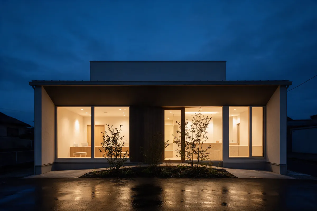 A small modern building at dusk, its windows lit and a few shrubs at the entrance